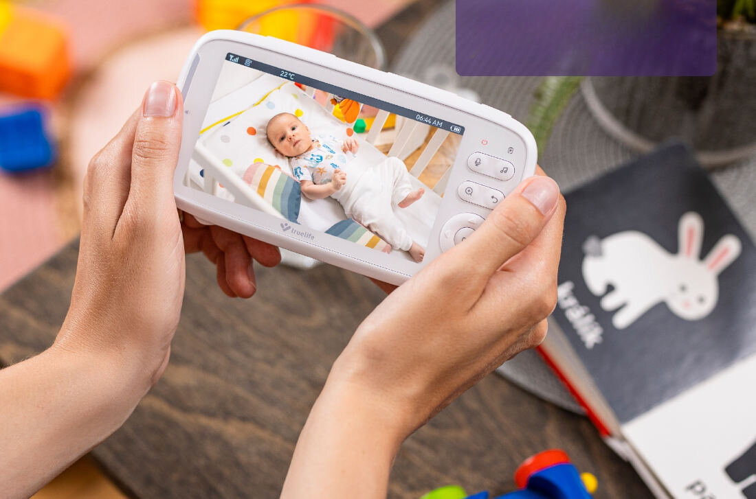 Osoba trzyma urządzenie z ekranem, na którym widać dziecko leżące na kolorowym kocyku. Na ekranie widoczny jest tekst: \'21°C\' oraz \'09:42 AM\'.