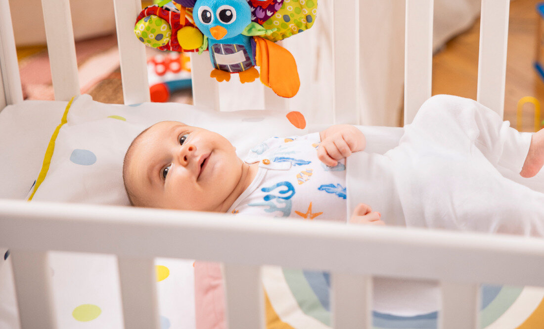 Niemowlę leży w łóżeczku, ubrany w biały śpioszek z kolorowymi wzorami. Nad nim wisi kolorowa zabawka w kształcie ptaka.