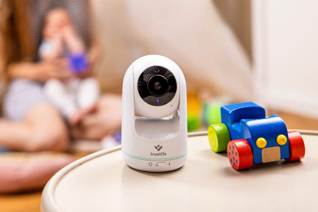 Na okrągłym stoliku znajduje się biała elektroniczna niania z logo \'truelife\' oraz kolorowa, drewniana zabawka w kształcie samochodu. W tle widoczna jest osoba trzymająca dziecko.
