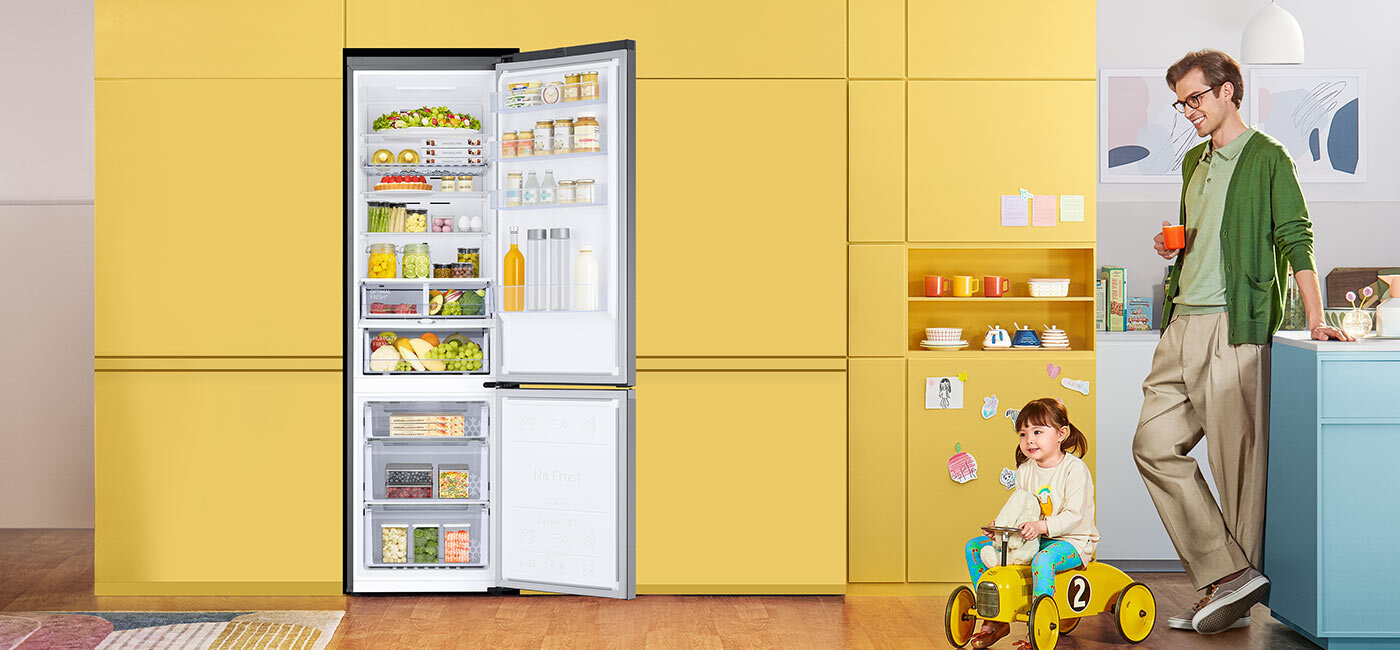 Sprawdź na zdjęciu, jak można układać produkty w chłodziarko-zamrażarce Samsung Lodówka z otwartymi drzwiami, wypełniona różnorodnymi produktami spożywczymi, stoi na tle żółtej ściany. Obok lodówki znajduje się dziecko na żółtym jeździku oraz osoba dorosła z kubkiem w ręku.