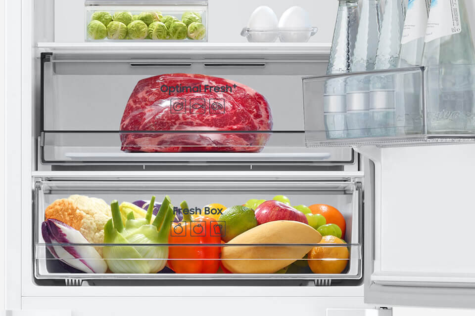 Wnętrze lodówki z widocznymi szufladami oznaczonymi jako \'Optimal Fresh\' i \'Fresh Box\', wypełnionymi mięsem oraz różnorodnymi warzywami i owocami. Na drzwiach lodówki znajdują się butelki z wodą, a na półce jajka i brukselka.
