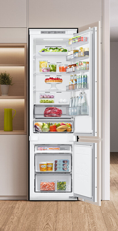 Otwarta lodówka w zabudowie, wypełniona różnorodnymi produktami spożywczymi, takimi jak warzywa, napoje, mięso i nabiał. Na drzwiach widoczne są półki z butelkami i kartonami, a w dolnej części znajdują się szuflady z mrożonkami.