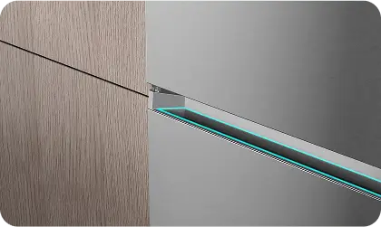 Zbliżenie na fragment lodówki z widocznym srebrnym panelem i drewnianą powierzchnią obok. Widać delikatne podświetlenie w kolorze niebieskim wzdłuż krawędzi panelu.