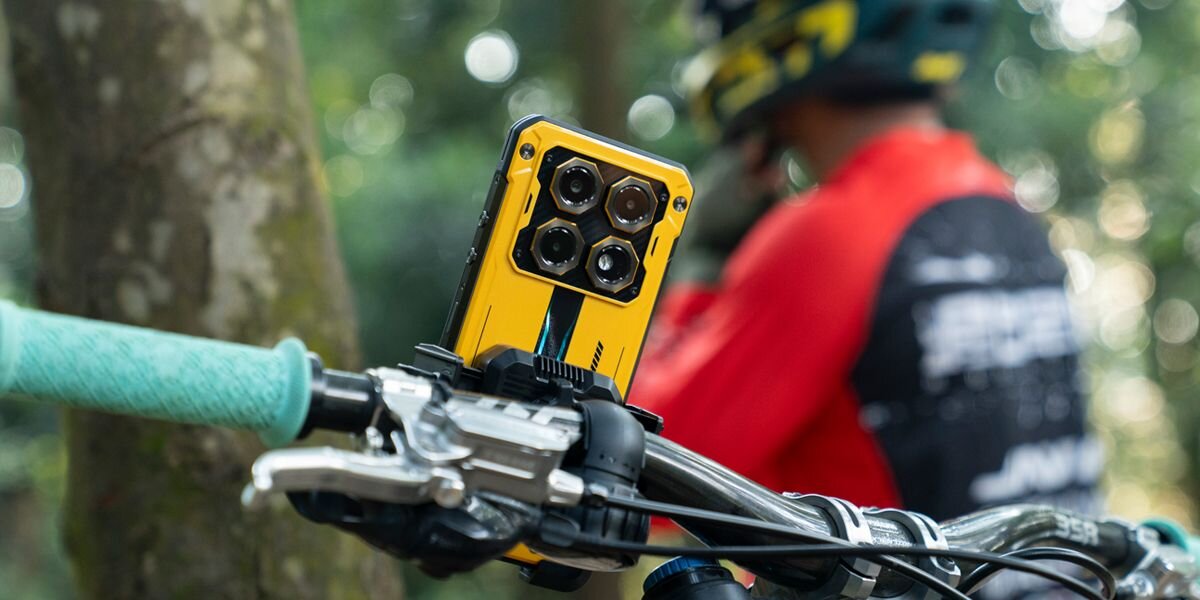 Żółty smartfon z czterema aparatami zamontowany na kierownicy roweru, w tle osoba w kasku i czerwonej odzieży sportowej.