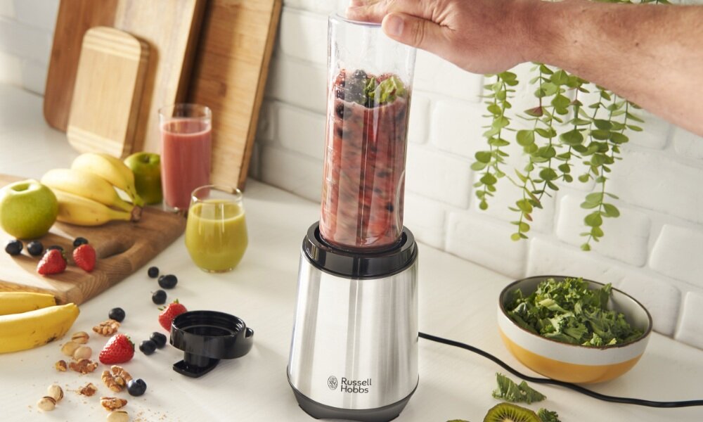 Na blacie kuchennym stoi blender z logo \'Russell Hobbs\', w którym znajduje się mieszanka owoców. Obok leżą owoce, orzechy i szklanki z sokami, a w tle widać deski do krojenia i roślinę doniczkową.