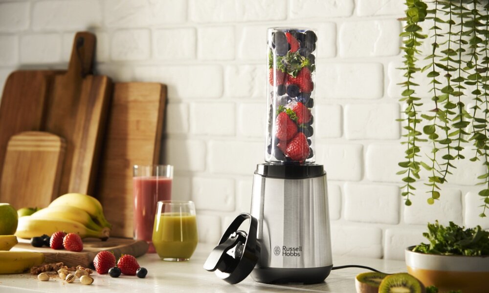 Na blacie kuchennym stoi blender z logo \'Russell Hobbs\', w którym znajduje się mieszanka owoców. W tle widoczne są deski do krojenia, owoce oraz szklanki z sokami.