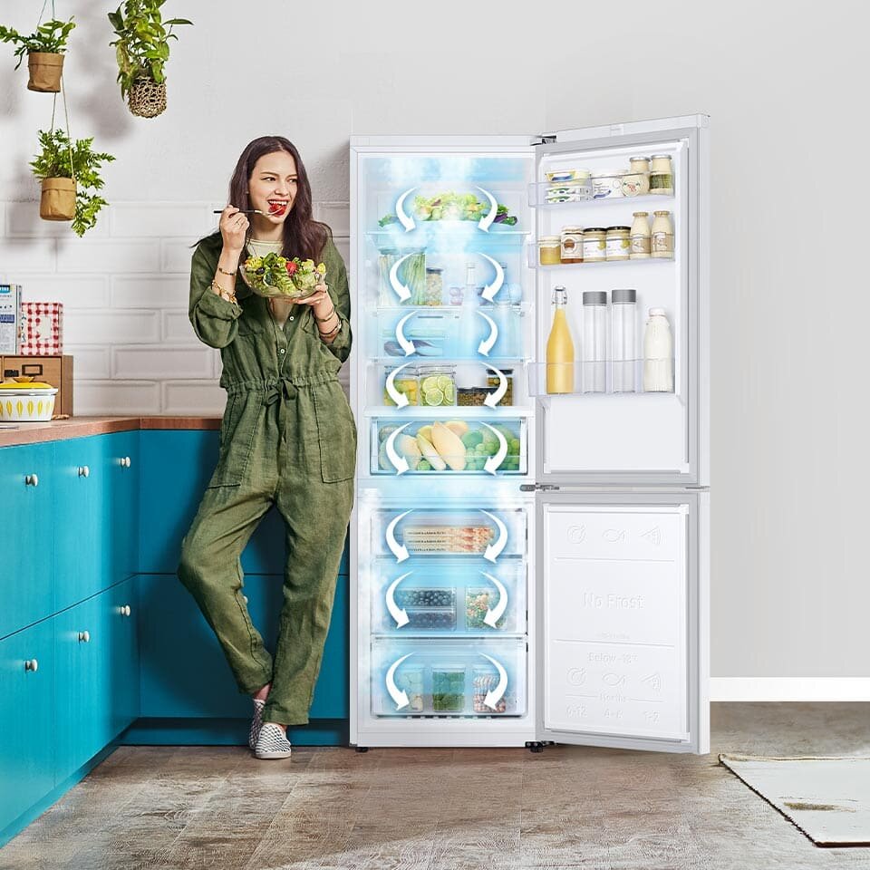Otwarta biała lodówka z widocznymi produktami spożywczymi na półkach i w drzwiach. Obok lodówki stoi osoba trzymająca miskę z sałatką.