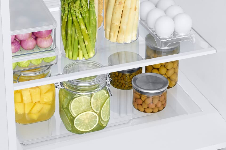 Wnętrze lodówki z półkami wypełnionymi różnymi produktami spożywczymi, takimi jak słoiki z limonkami i oliwkami, pojemniki z szparagami oraz jajka w plastikowym opakowaniu. Produkty są starannie ułożone na szklanych półkach.