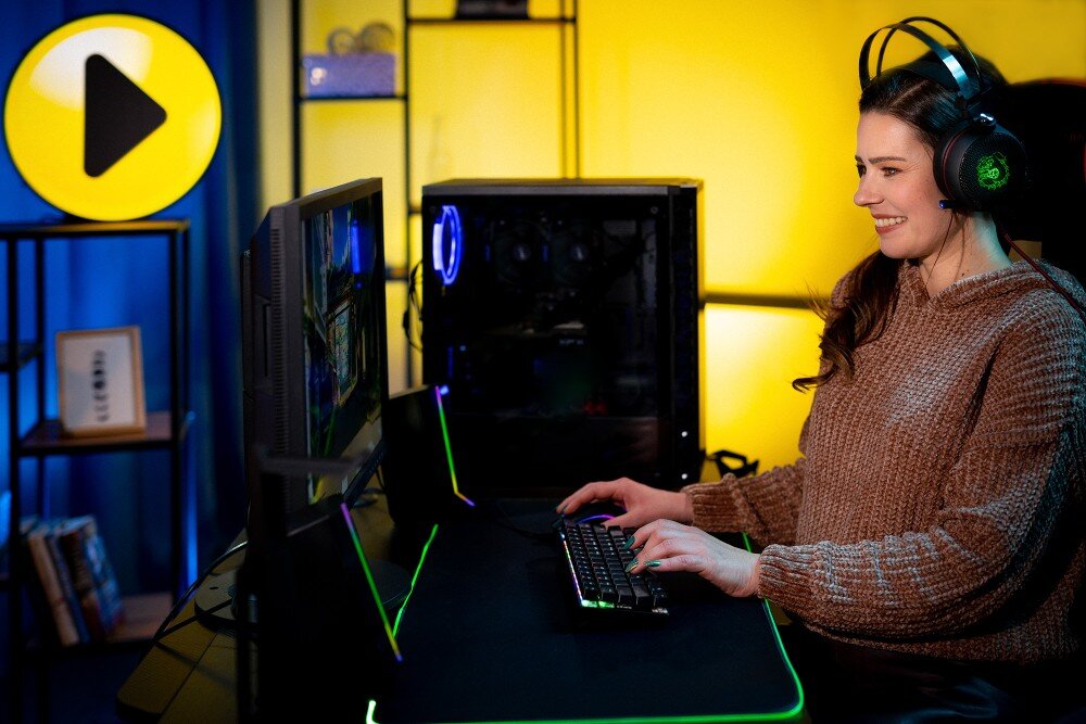 Komputer MAD DOG MD-A07DR16 Kobieta w brązowym swetrze i słuchawkach siedzi przed komputerem i gra w grę na monitorze w oświetlonym pokoju