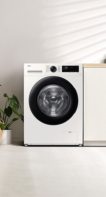 Biała pralka z czarnym okrągłym bębnem i panelem sterowania z wyświetlaczem cyfrowym. Obok pralki stoi roślina w doniczce na jasnej podłodze.