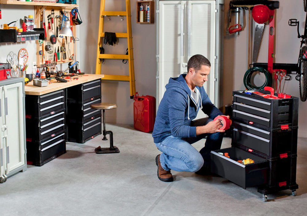 Regał z szufladami KETER 17201228 Czarny pomieszczenie garaż warsztat regał wypełniony narzędziami i różnymi rzeczami stoi na podłodze osoba klęczy przy regale z taśmą pojemny regał porządek przechowywanie transport mobilność