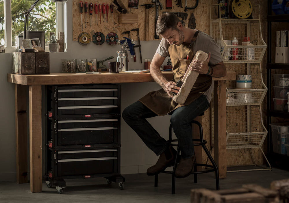 Regał z szufladami KETER 17201228 Czarny pomieszczenie pracowania stolarska regał umieszczony pod stołem roboczym obok siedzi na krześle osoba z kawałkiem drewna w dłoni w tle ściana wypełniona narzędziami okno pojemność do 50,5 l wymiary 28,9 x 56,2 x 74,2 cm optymalne rozplanowanie szuflad i pojemników szybki dostęp do narzędzi