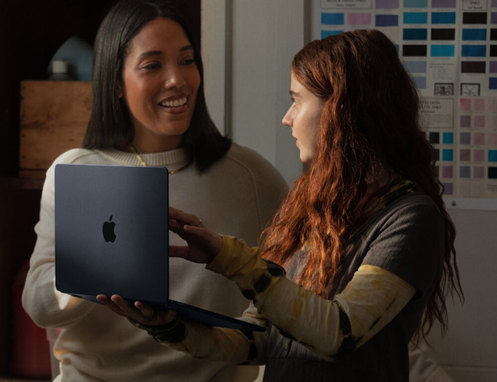 Dwie osoby trzymają laptopa z widocznym logo w kształcie nadgryzionego jabłka. W tle znajduje się tablica z próbkami kolorów.