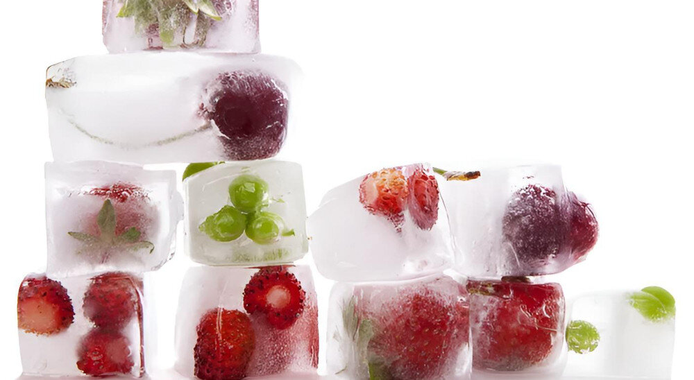 Na obrazie widoczne są kostki lodu z zamrożonymi owocami, takimi jak truskawki, winogrona i wiśnie. Kostki są ułożone jedna na drugiej, tworząc nieregularną strukturę.