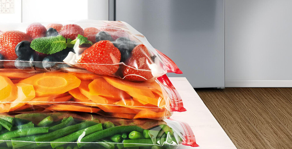 Na blacie leżą trzy przezroczyste worki z zamrożonymi owocami i warzywami: truskawkami, marchewką i fasolką szparagową. W tle widoczna jest stalowa lodówka.