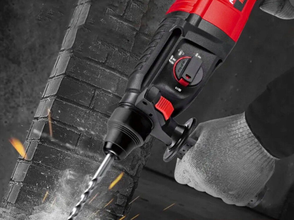 Osoba w rękawicy trzyma młotowiertarkę, która wierci w ceglanej ścianie, generując iskry. Urządzenie jest czarno-czerwone z widocznymi pokrętłami i przyciskiem.