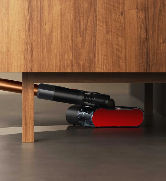 Elektryczny mop z czerwonym elementem czyszczącym znajduje się pod drewnianym meblem. Urządzenie ma czarną obudowę i miedziany uchwyt.