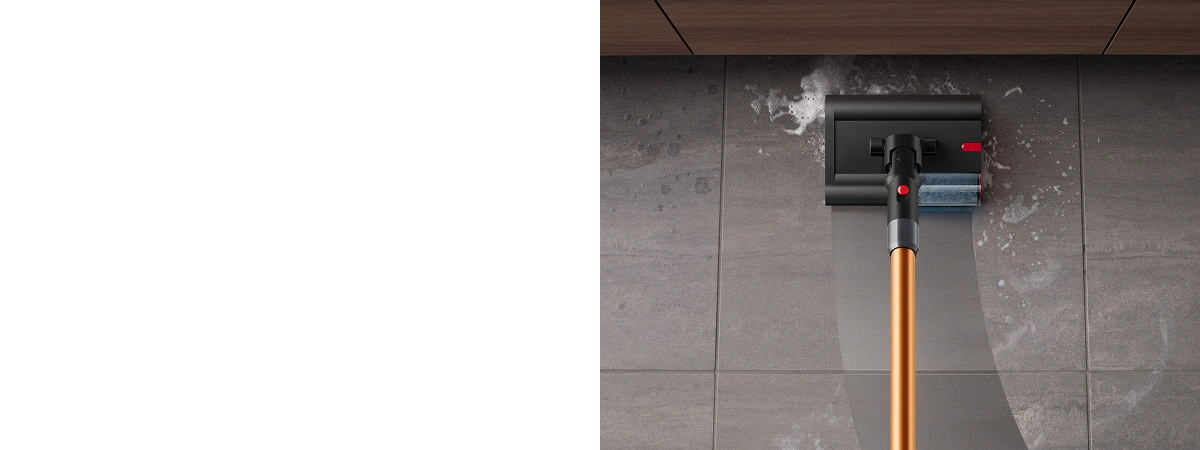 Na obrazie widać mop elektryczny z miedzianą rurą, czyszczący mokrą podłogę z ciemnych płytek. Mop ma czarną głowicę z niebieską szczotką i czerwonym przyciskiem.