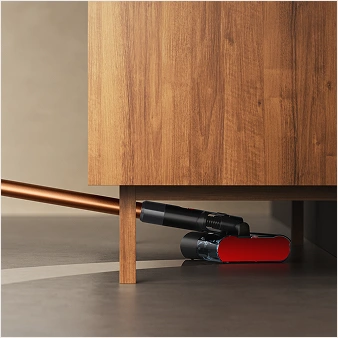 Elektryczny mop z czerwonym elementem czyszczącym znajduje się pod drewnianą szafką na gładkiej podłodze.