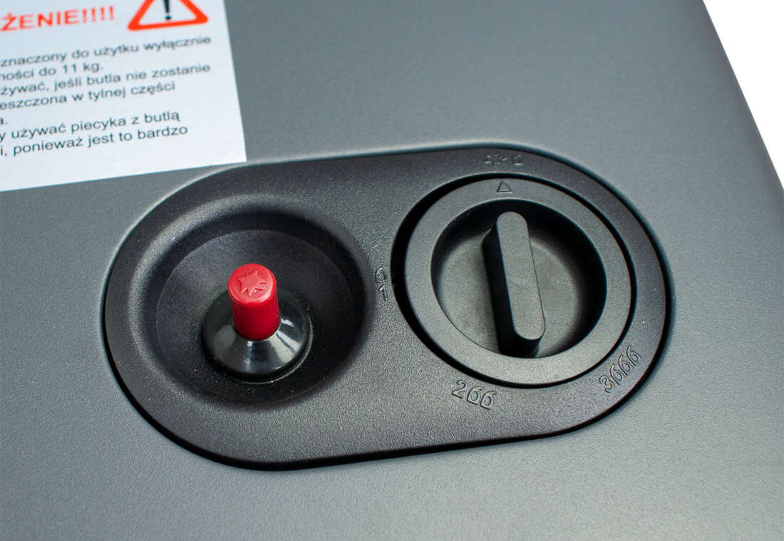 Zdjęcie przedstawia panel sterowania piecyka gazowego z czerwonym przyciskiem i pokrętłem. Obok znajduje się ostrzeżenie z tekstem: \'PRZEZNACZONY DO UŻYTKU WYŁĄCZNIE\'.