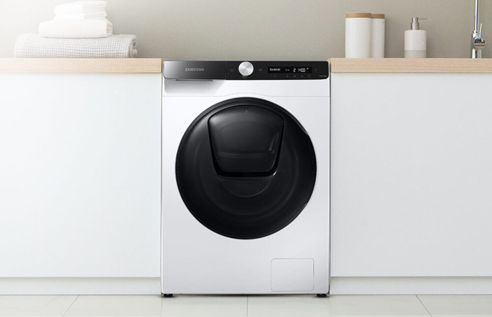 Pralko-suszarka w białym kolorze z czarnym, okrągłym panelem drzwiowym, umieszczona pomiędzy białymi szafkami. Na górze urządzenia widoczne jest logo \'Samsung\' oraz panel sterowania z pokrętłem i wyświetlaczem.