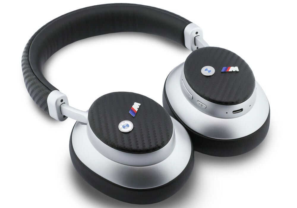 Srebrne słuchawki nauszne z czarnymi elementami o fakturze włókna węglowego, z widocznym logo BMW M. Na jednej z muszli znajdują się przyciski oraz port ładowania.