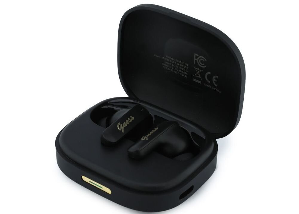 Czarne słuchawki douszne z logo \'Guess\' w złotym kolorze znajdują się w otwartym etui ładującym. Wewnątrz pokrywy etui widoczne są oznaczenia certyfikacyjne oraz napis \'Made in China\'.