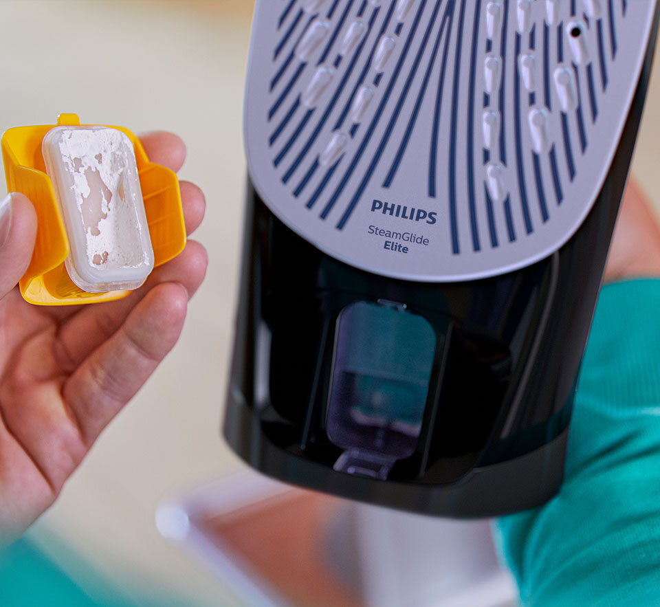 Osoba trzyma żółty pojemnik z białym osadem obok żelazka z widoczną stopą SteamGlide Elite i logo PHILIPS.