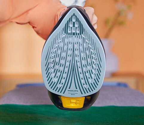 Zdjęcie przedstawia stopę żelazka z widocznymi otworami parowymi i napisem \'PHILIPS Azur 7000 Series\'. W tle rozmyty widok wnętrza z rośliną.