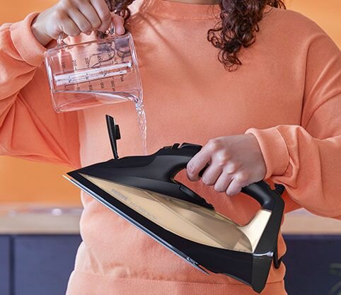 Osoba trzyma żelazko i nalewa do niego wodę z miarki. Żelazko ma czarną obudowę i metaliczną stopę.