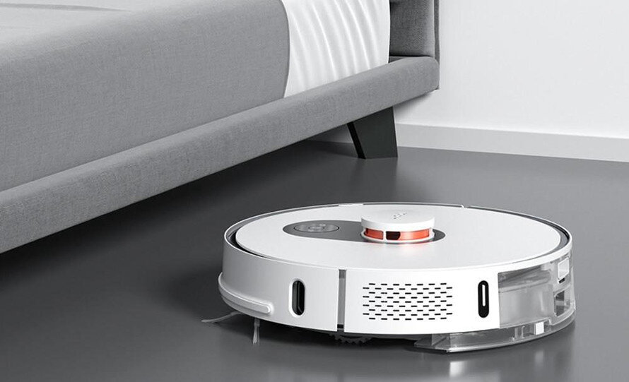 Na obrazie widoczny jest biały robot sprzątający na podłodze obok łóżka. Robot ma okrągły kształt i widoczne są na nim otwory wentylacyjne oraz przyciski sterujące.