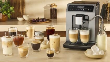 Ekspres do kawy KRUPS z dwoma szklankami przy dozowniku stoi na blacie, obok kilka szklanek z różną kawą.