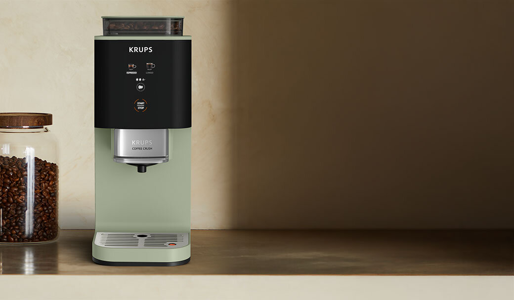Ekspres do kawy marki KRUPS z napisem \'COFFEE CRUSH\' na przednim panelu, obok słoik z ziarnami kawy.