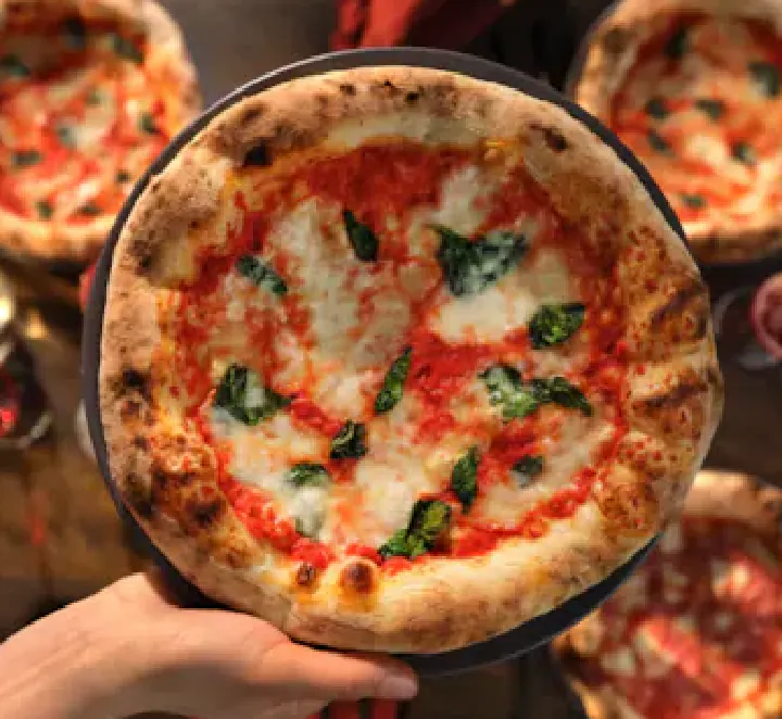 Na obrazie widoczna jest pizza z sosem pomidorowym, roztopionym serem i liśćmi bazylii. Pizza jest trzymana na talerzu, a w tle widać inne pizze na stole.