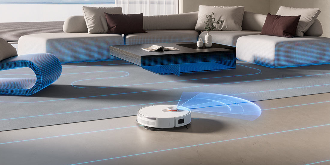Biały robot sprzątający porusza się po podłodze w nowoczesnym salonie z jasnymi meblami. Na podłodze widoczne są niebieskie linie wskazujące trasę robota.