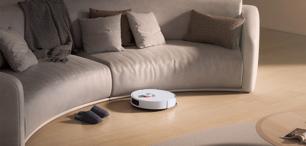 Na drewnianej podłodze w salonie znajduje się biały robot sprzątający. W tle widoczna jest jasna, zakrzywiona sofa z kilkoma poduszkami.