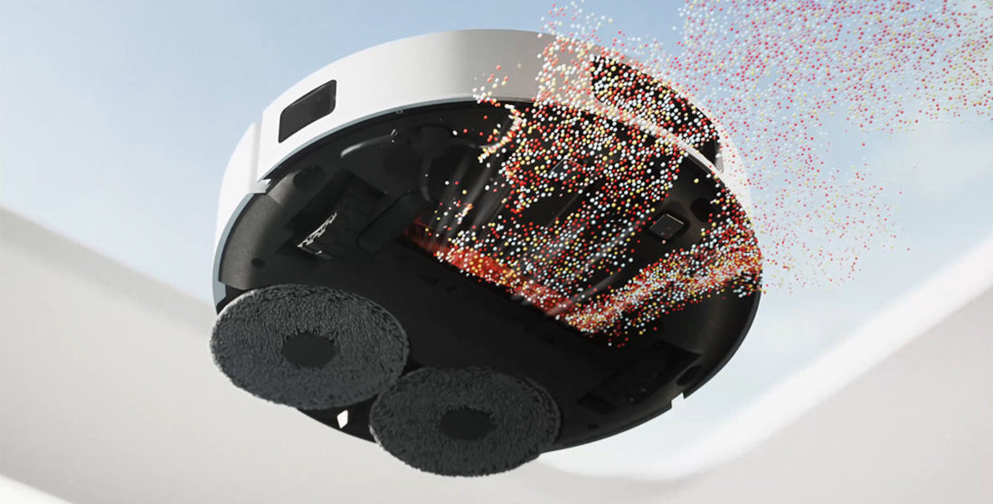 Obraz przedstawia biały robot sprzątający z widocznymi dwoma okrągłymi szczotkami od spodu. Z urządzenia wydobywają się kolorowe cząsteczki, sugerujące działanie ssące lub czyszczące.