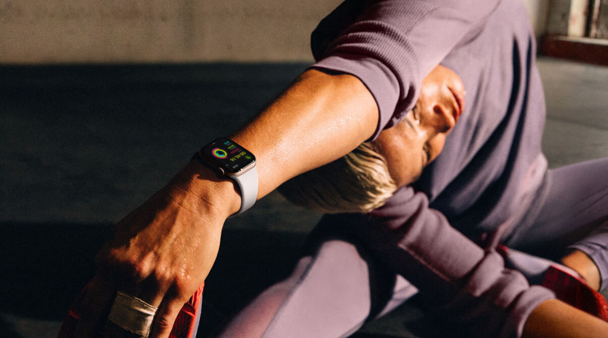 Osoba w sportowym stroju wykonuje ćwiczenie rozciągające, nosząc smartwatch z jasną opaską. Na ekranie zegarka widoczne są kolorowe pierścienie aktywności.