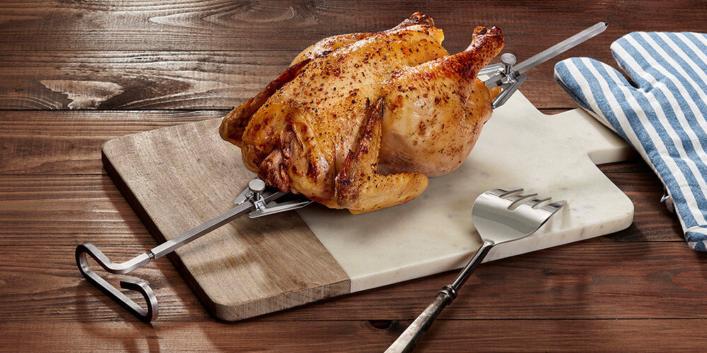 Na drewnianej desce do krojenia leży upieczony kurczak na rożnie. Obok znajduje się metalowy widelec do mięsa i rękawica kuchenna w biało-niebieskie paski.