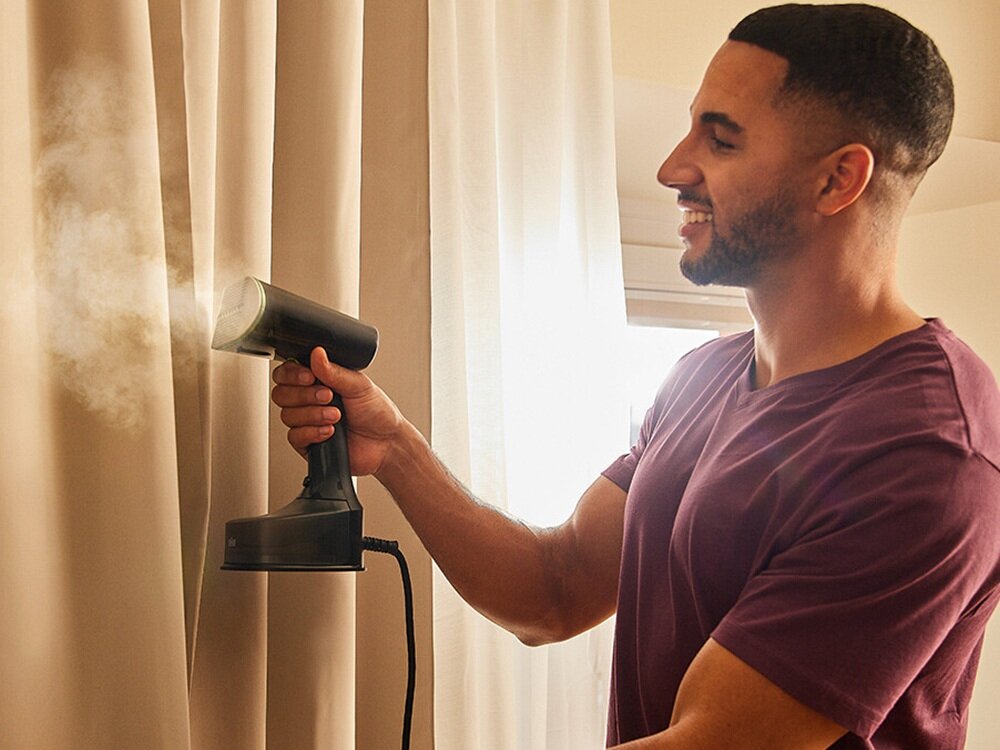 Osoba używa parownicy do ubrań, z której wydobywa się para, na tle zasłon. Parownica jest koloru czarnego i ma nowoczesny design.