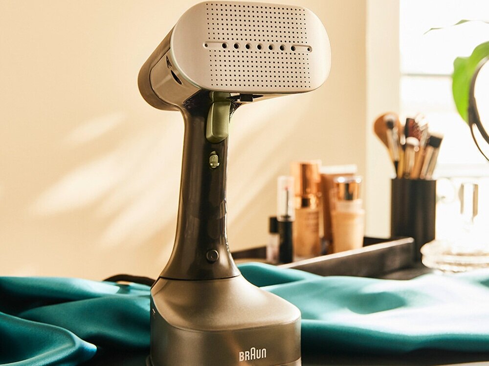 Na zdjęciu znajduje się parownica do ubrań marki Braun, ustawiona na stole obok kosmetyków. Na urządzeniu widoczne jest logo \'BRAUN\'.