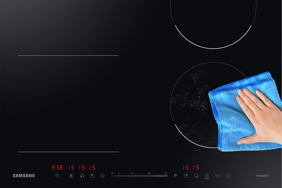 Czarna płyta indukcyjna z widocznym logo SAMSUNG i cyfrowym panelem sterowania z czerwonymi cyframi. Na płycie widoczna jest ręka trzymająca niebieską ściereczkę, czyszcząca powierzchnię.
