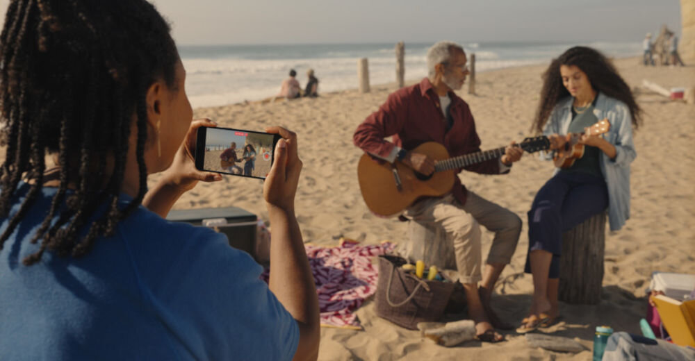 Osoba trzyma smartfon i nagrywa dwie osoby grające na instrumentach na plaży. W tle widać morze i kilka osób siedzących na piasku.