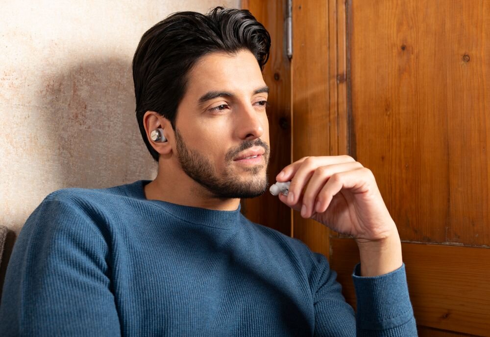 Osoba nosi słuchawki dokanałowe w kolorze czarnym, siedząc przy drewnianej ścianie. Ubrana jest w niebieski sweter.