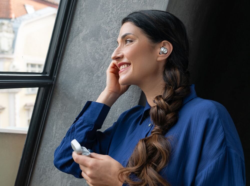 Osoba w niebieskiej koszuli stoi przy oknie, nosząc słuchawki dokanałowe. W ręku trzyma etui na słuchawki.