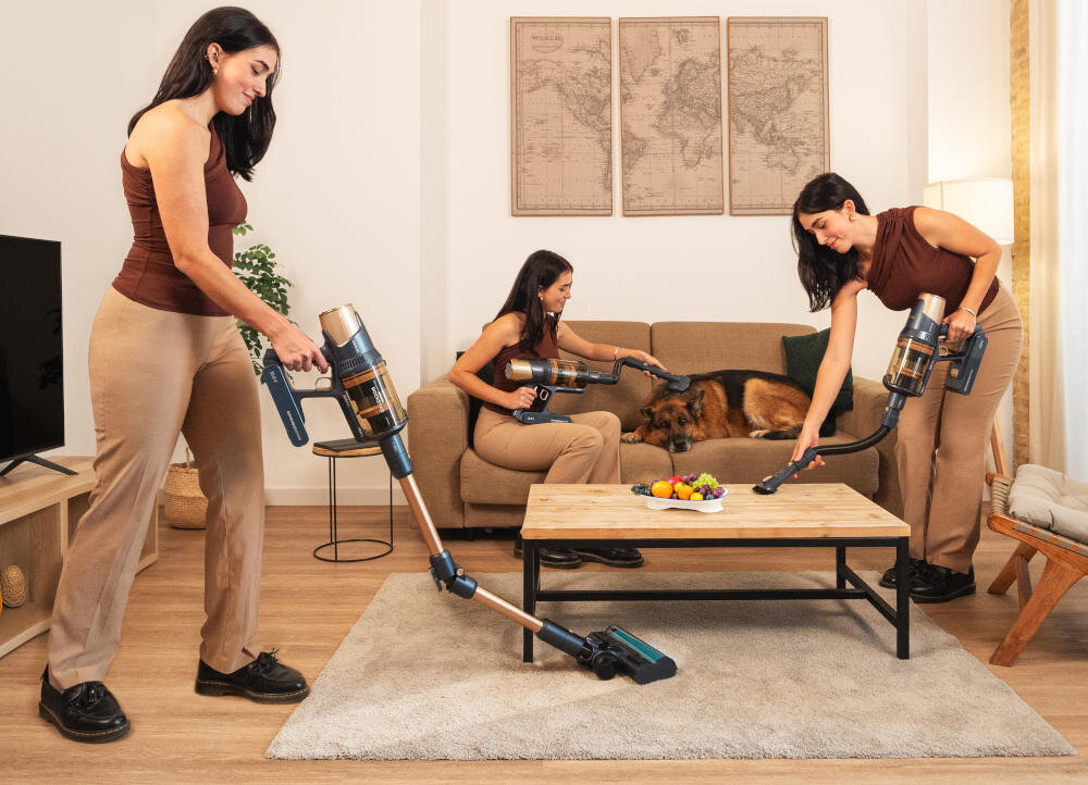Trzy kobiety używają odkurzacza bezprzewodowego w salonie, w którym znajduje się kanapa, stolik z owocami i dywan. Na kanapie leży pies, a na ścianie wiszą mapy.
