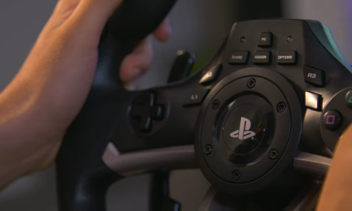 Zbliżenie na kierownicę do gier z widocznym logo PlayStation na środku. Wokół znajdują się przyciski funkcyjne, takie jak SHARE, ASSIGN, OPTIONS oraz symbole charakterystyczne dla kontrolera PlayStation.