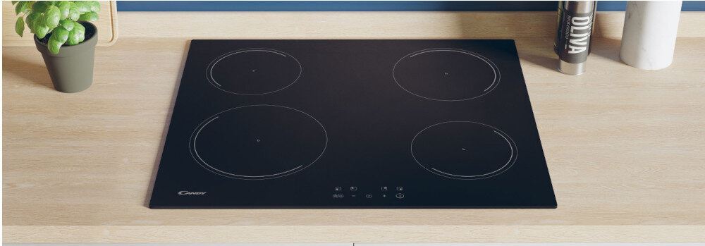 Czarna płyta indukcyjna z czterema okrągłymi polami grzewczymi, umieszczona na drewnianym blacie. Obok znajduje się zielona roślina w doniczce oraz pojemnik z napisem \'OLIVA\'.