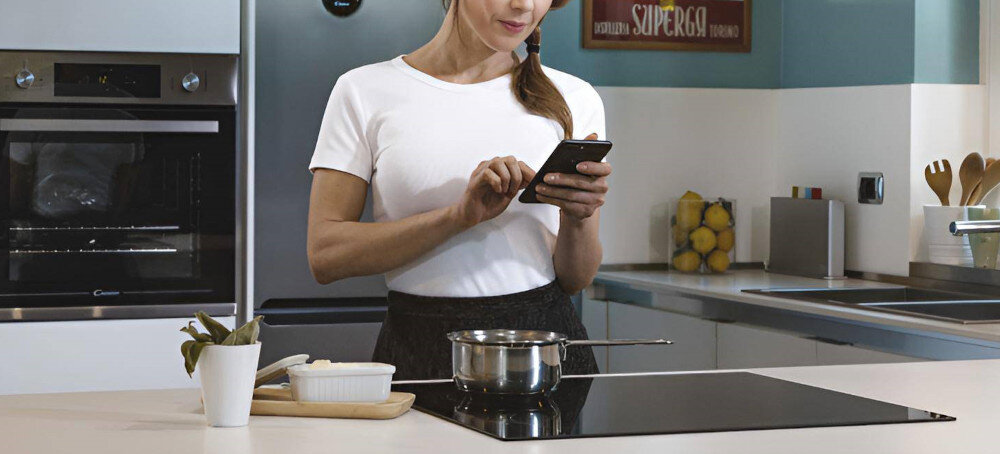 Kobieta stoi przy kuchennym blacie, na którym znajduje się płyta indukcyjna z garnkiem. W tle widoczny jest piekarnik oraz lodówka, a na blacie leży deska z naczyniem i kubkiem.