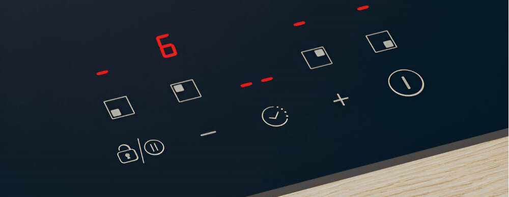 Panel sterowania płyty indukcyjnej z wyświetlaczem pokazującym cyfrę 6 w kolorze czerwonym. Widoczne są przyciski dotykowe z symbolami, takimi jak blokada, pauza, zegar, plus i minus.